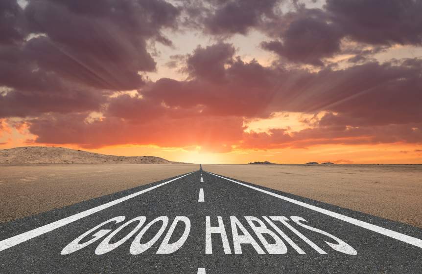 A long straight road in the middle of a barren desert leading off into a very colourful sunset. At the start of the road are the words "good habits" painted on it.