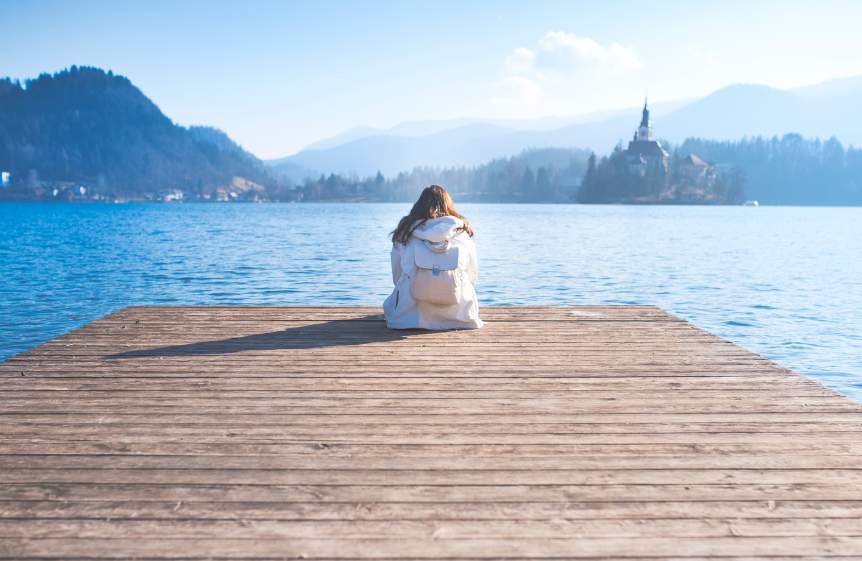 A girl in a white jacket with a white bag on her back looking out over the water looking for her clarity of purpose. In the distance is hills, mountains trees and a church on an island.
